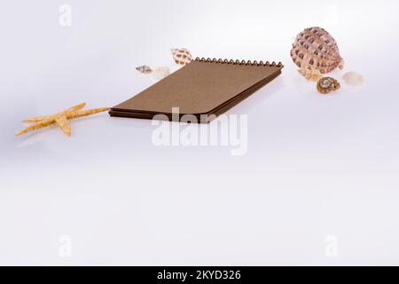 Kleines braunes Spiralheft mit Muscheln auf weißem Hintergrund Stockfoto