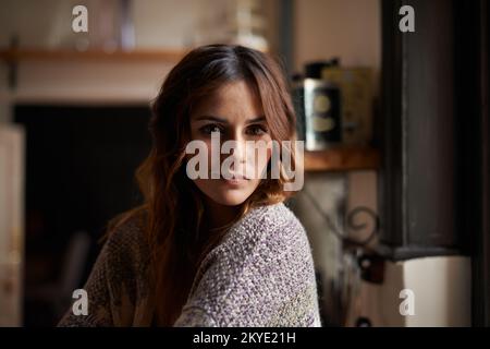 Ruhige Morgenstunden...Porträt einer wunderschönen jungen brünetten Frau, die in einer Küche sitzt und in Fensterlicht getaucht ist. Stockfoto