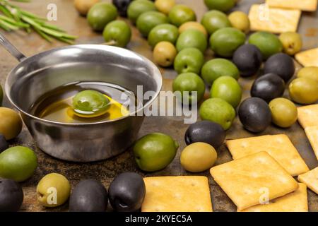 Schwarze und grüne Oliven, Kekse auf dem Tisch. Olivenöl in einer Schüssel. Schließen. Rostiger Metallhintergrund. Stockfoto