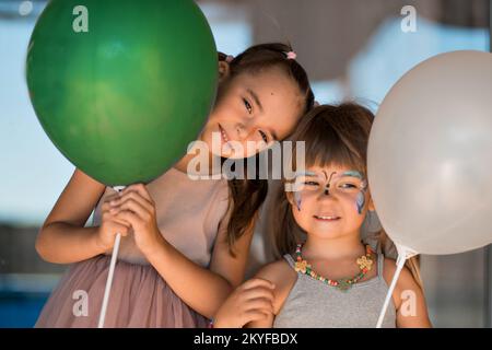 Zwei kleine Schwestern mit Gesichtsmalerei lächeln mit Ballons. Kinderurlaub, glückliche Kinder. Das Mädchen hat Geburtstag. Hochwertiges Foto. Stockfoto