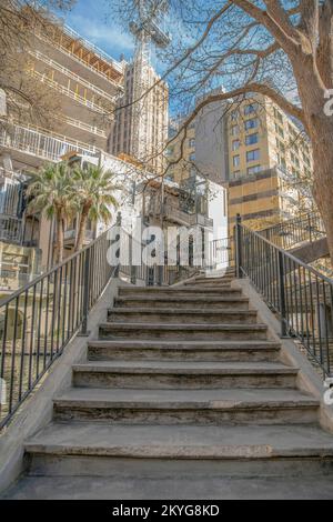 Treppen mit Geländern, die eine Brücke im San Antonio River Walk in Texas hinaufführen. Malerische Ausblicke auf Gebäude entlang des Kanalwassers können Sie im Hinterland sehen Stockfoto