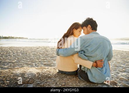 Warten auf den Sonnenuntergang...Rückblick auf ein junges Paar, das am Strand saß und liebevoll war. Stockfoto