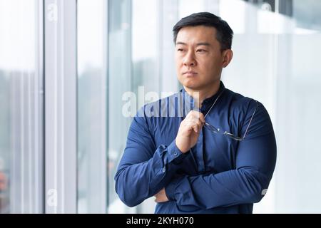 Asiatischer Geschäftsmann, Mann mit Hemd und Brille am Bürofenster, seriöser Boss, der über zukünftige Pläne und Strategien nachdenkt. Stockfoto