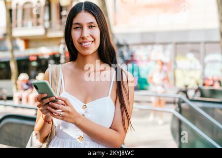 Optimistisch hispanische Frau in weißem Kleid mit Rucksack, die ihr Smartphone durchsucht und mit einem Lächeln auf die Kamera schaut, auf einem unscharfen Hintergrund der sonnenbeleuchteten Straße in Barcelona, Spanien Stockfoto