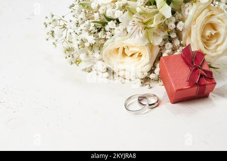 Zwei Ringe, rote Geschenkbox, Blumenstrauß auf weißem Holzhintergrund. Wird in der oberen rechten Ecke des Fotos platziert. Stockfoto