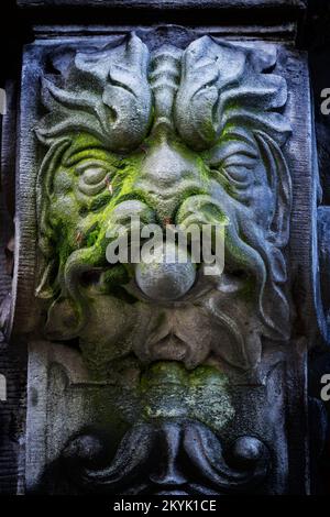 Monstergesicht mit Löwenmähne und Schnurrbart, gruselige verwitterte Steinskulptur mit Moos, Fantasy Kreatur Steinwerk Dekoration auf einer Veranda Stockfoto