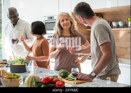 Group of diverse middle aged friends chatting in the kitchen while preparing food Stock Photo