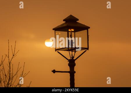 Malvern Wells, Worcestershire, Großbritannien. 1.. Dezember 2022. Die Sonne geht heute Morgen durch Nebel hinter einer der wenigen Gaslampen des Landes in Malvern Wells, Worcestershire, auf. Die Straßenbeleuchtung mit Gaslampen endete Ende der 1950er Jahre nach der Umwandlung in Elektrizität. Great Malvern ist eine von wenigen Städten, in denen noch einige Gaslampen verwendet werden, darunter Westminster. Kredit: Peter Lopeman/Alamy Live News Stockfoto