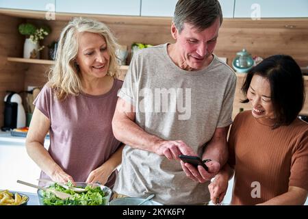 Älterer Mann, der ein Smartphone benutzt, während er mit Freunden in der Küche spricht Stockfoto