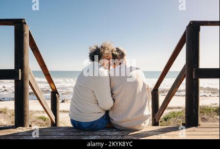 Romantische Seniorin, die glücklich lächelt, während sie mit ihrem Mann auf einer hölzernen Brücke am Meer sitzt. Ein älteres Paar im Ruhestand verbringt etwas Zeit Stockfoto