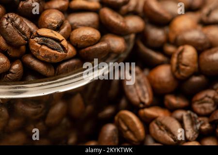 Geröstete Kaffeebohnen auf dem Tisch verstreut und eine kleine Kaffeetasse. Nahaufnahme, selektiver Fokus. Stockfoto