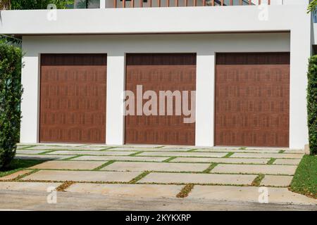 Drei dunkle Holzgaragentore mit Graspflastereinfahrt in Miami, Florida. Garagenaußenseite mit bemalten weißen Wänden unter einer Terrasse. Stockfoto