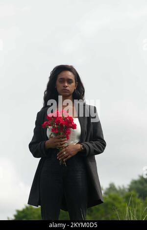 Junge schwarze Frau, die einen Haufen roter Blumen hält, die melancholisch aussehen. Er trägt ein weißes Hemd und einen dunklen Mantel. Stockfoto