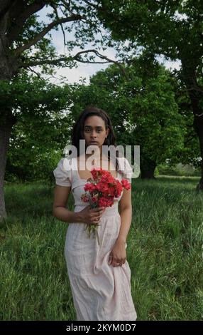 Junge schwarze Frau in weißem Kleid und mit roten Blumen in Eichenwäldern. Stockfoto