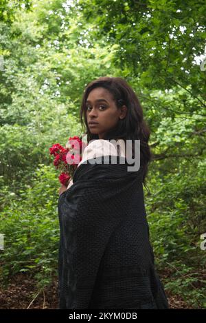 Eine junge Frau im Wald, die ein weißes Kleid mit schwarzem Schal trägt und einen Haufen roter Blumen in der Hand hat. Stockfoto