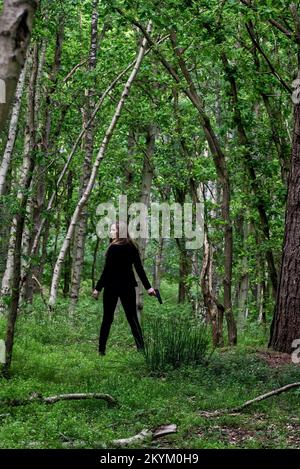 Rothaarige junge Frau in Schwarz und mit einer Pistole im Wald, Thriller, Mystery, Adventure Book Cover Style. Stockfoto