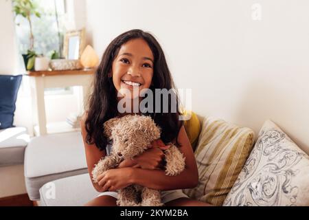 Porträt eines glücklichen Mädchens, das auf einem Sofa im Wohnzimmer sitzt und Teddybär umarmt Stockfoto