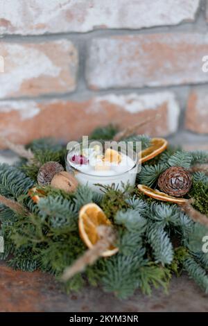 Dekorative Kerze mit Christbaumzweigen, getrockneten Orangen, Nüssen und Zapfen auf Backsteinwänden Stockfoto