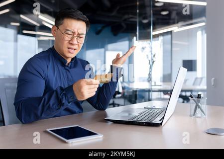 Frustrierter asiatischer Mann, der versuchte, Banktransaktionen und Einkäufe im Online-Shop zu tätigen, ein Geschäftsmann betrog und traurig, als er im Büro mit einem Laptop und einer Bankkreditkarte arbeitete. Stockfoto