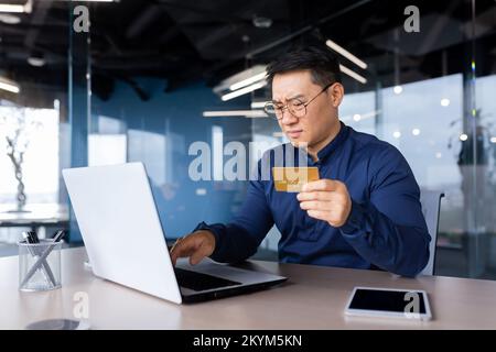 Frustrierter asiatischer Mann, der versuchte, Banktransaktionen und Einkäufe im Online-Shop zu tätigen, ein Geschäftsmann betrog und traurig, als er im Büro mit einem Laptop und einer Bankkreditkarte arbeitete. Stockfoto