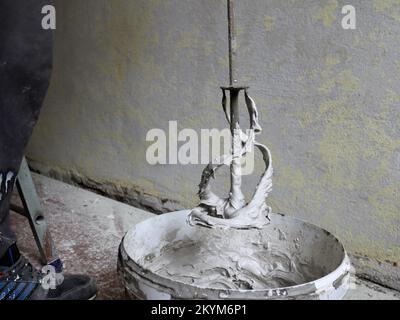 Ein Schneebesen zum Mörtelmischen, der über einem weißen Kunststoffeimer in einem Raum mit alten verfallenen Wänden, einem Reparaturarbeitsplatz und einem Gipswerkzeug gehoben wird Stockfoto