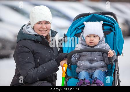 Mutter in warmer Winterjacke und Hut geht mit einem zweijährigen Kind im Kinderwagen Stockfoto