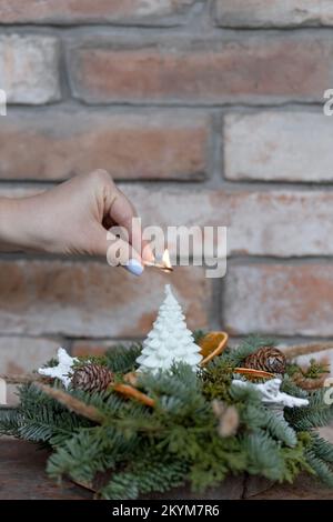 Die Hand einer Frau zündet eine Kerze in Form eines Weihnachtsbaums an, um die Weihnachtsgegenstände vor einem Backsteingebäude liegen Stockfoto