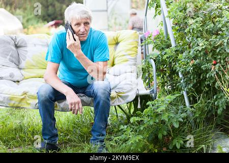 Ein älterer Mann sitzt auf einem Hängesessel und telefoniert im Hinterhof des Landhauses Stockfoto