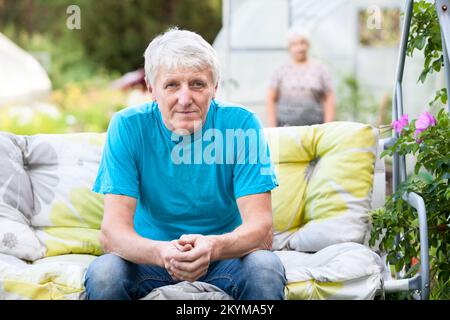 Ein Mann sitzt in einem Hängestuhl vor dem Hintergrund seiner Frau, die in der Nähe des Landhauses steht Stockfoto