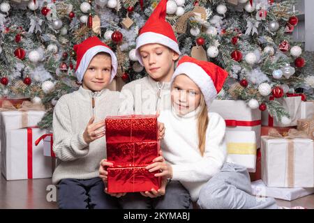 Drei süße, fröhliche, aufgeregte Kinder, Jungen und ein Mädchen mit weihnachtsmannmützen tauschen zu Hause Überraschungsgeschenke neben dem weihnachtsbaum aus. Schwester und Brüder geben Chri Stockfoto