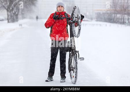 Ein Mann in einer roten Jacke hob ein Fahrrad hinter dem Lenkrad am Hinterrad, ein Winterschneetag Stockfoto