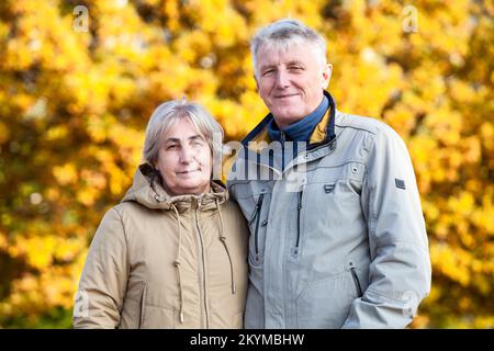 Ältere Ehepaare, die zusammen standen, eine Jacke gegen gelbe Herbstblätter von Bäumen trugen, auf die Kamera starrten und lächelnde Menschen anschauten Stockfoto