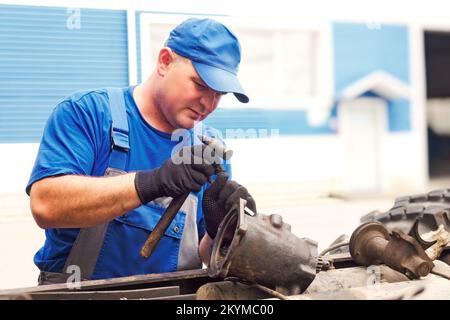 Mechaniker in Overalls und Baseballmütze repariert Traktor oder LKW am Sommertag. Vorderansicht. Professionelle Unterstützung bei der Wartung von schweren Geräten. Authentischer Workflow... Stockfoto