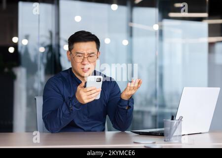 Ein Geschäftsmann liest online schlechte Nachrichten vom Telefon, ein asiatischer Mann ist enttäuscht und traurig, wenn er auf den Smartphone-Bildschirm schaut, ein Mann im Hemd, der im Büro mit einem Laptop auf der Arbeit arbeitet. Stockfoto