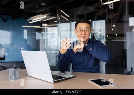 Asiatischer Geschäftsmann verwendet Smartwatch, um zu kommunizieren, der Mensch gibt Befehle an künstliche Intelligenz mit einer Armbanduhr, der Boss im Hemd arbeitet im Büro mit einem Laptop. Stockfoto