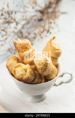 Hausgemachte Gebäckröhrchen mit Schlagsahne in der Tasse mit weißem Tee. Selektiver Fokus auf die nächstgelegene Süßröhre mit Schlagsahne. Schließen Stockfoto