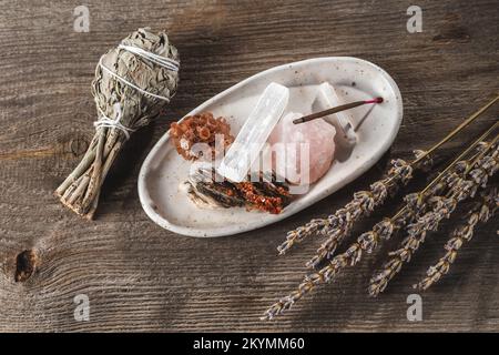 Weißer Keramikhalter mit mineralischen Kristallsteinen und Aroma-Stick. Ein Bündel Salvia Sage und Lavendel im rustikalen Holzhintergrund Stockfoto