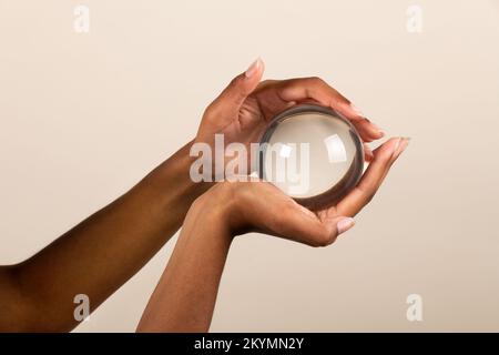 Hände einer schwarzen Frau, die eine transparente Kristallkugel mit Lichtreflexion auf beigefarbenem Hintergrund hält Stockfoto