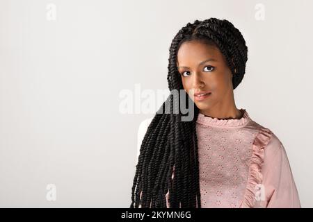 Sanfte junge afroamerikanische Frau in bescheidenem rosa Kleid, die Haare in stilvollen langen Afro-Geflechten hat und auf grauen Hintergrund auf die Kamera schaut Stockfoto