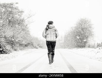 Schwarz-weiße Rückansicht einer Frau, in Wintermantel und Gummistiefeln, die mitten auf einer ländlichen Landstraße im Schnee isoliert läuft. Stockfoto
