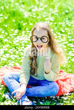 Das Mädchen sitzt auf Gras am Grassplot, grüner Hintergrund. Kinder posieren mit einer Pappbrille für eine Fotosession auf der Wiese. Intelligentes und intelligentes Konzept. Mädchen Stockfoto