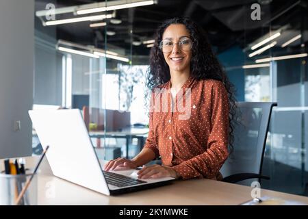 Junge, wunderschöne hispanische Frau, die in einem modernen Büro arbeitet, lächelt und mit einem Laptop in die Kamera schaut. Stockfoto