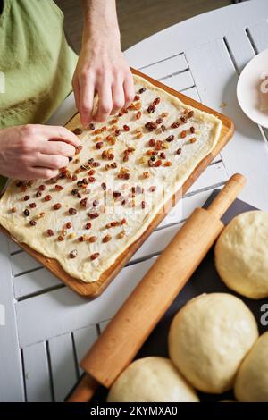 Ostergebäck, Cruffin, Zubereitung zu Hause Konzept. Eine nicht erkennbare Person streut Rosinen auf mit Butter bestrichenen Teig. Männchen gießt Rosinen auf einen Teig mit Butter. Hochwertiges Foto Stockfoto