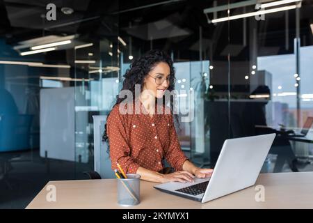 Fröhliche und erfolgreiche indische Programmiererin bei der Arbeit in modernen Büros, technische Supportmitarbeiterin mit lächelndem Laptop-Tippen auf der Tastatur. Stockfoto