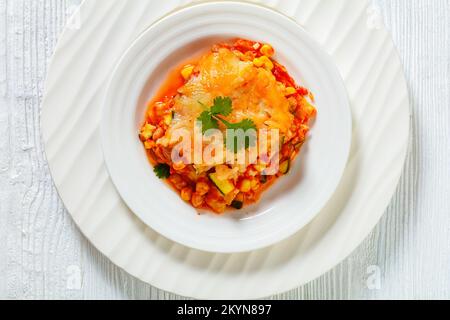 Vegetarischer Enchilada-Auflauf mit Tortillas, Sauce, Pintobohnen, Mais, Zucchini und mexikanischem Mischkäse auf weißem Teller, Nahaufnahme Stockfoto