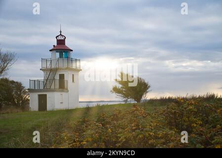 Leuchtturm, Spodsbjerg Fyr in Huntsted an der Küste Dänemarks. Die Sonne scheint durch die Wolken. Wiese mit Bäumen. Landschaftsfoto vom Meer Stockfoto