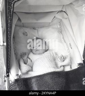 1950er, historisch, Blick von oben auf ein Baby, das auf dem Rücken eines weichen Kissens in einem Kinderwagen liegt, mit einer hellen Baumwolldecke und einem Klappdach des Kinderwagens, England, Großbritannien. Stockfoto