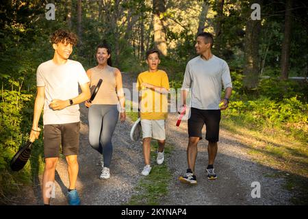 Glückliche Familie mit Tennisball und Schlägern, die während des Urlaubs auf einer Feldstraße im Wald spazieren Stockfoto