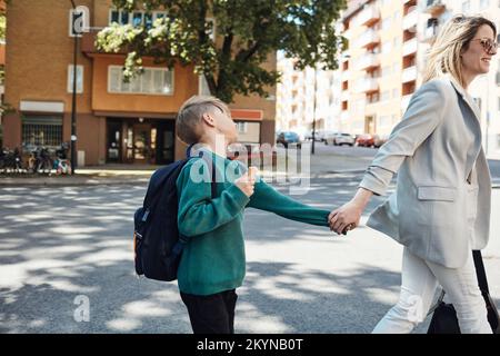 Seitenansicht einer Frau, die einen sturen Sohn zur Schule bringt, während sie auf der Straße spaziert Stockfoto