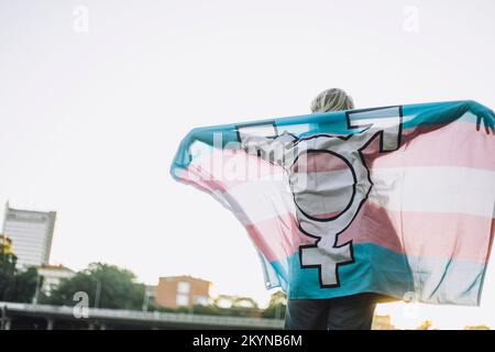 Rückansicht einer nicht-binären Person, die mit LGBTQIA-Flagge am klaren Himmel steht Stockfoto